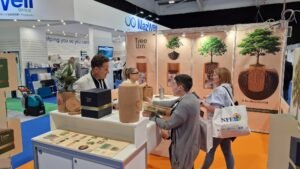 Exhibition stand with orange carpet, four. People chatting around a white piece of furniture or on which their eyes are fixed, Biodegradable Funerary, blue packaging and a green plant, children, another stand, white and blue and a floor washing machine.